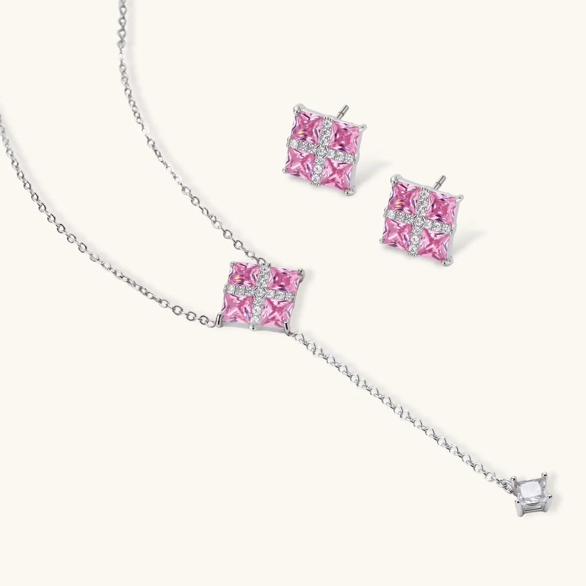 Necklace and stud earrings set with four pink square zirconia stones and a white crossed band, on a cream background.