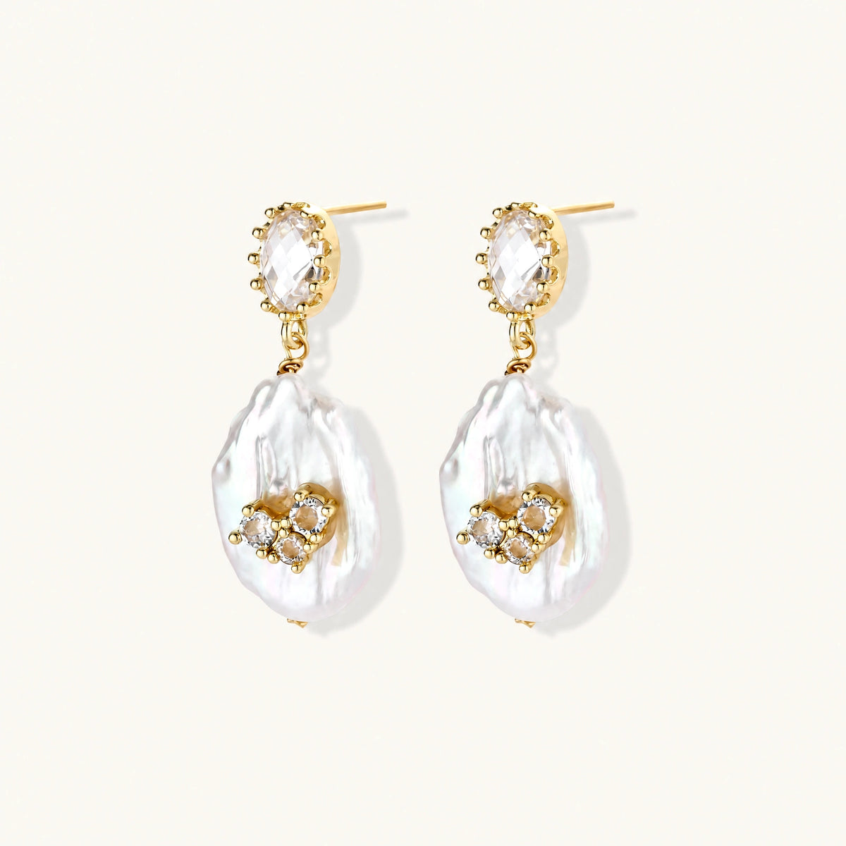 Gold-plated drop earrings with iridescent baroque pearls and crystal flower accents on cream background