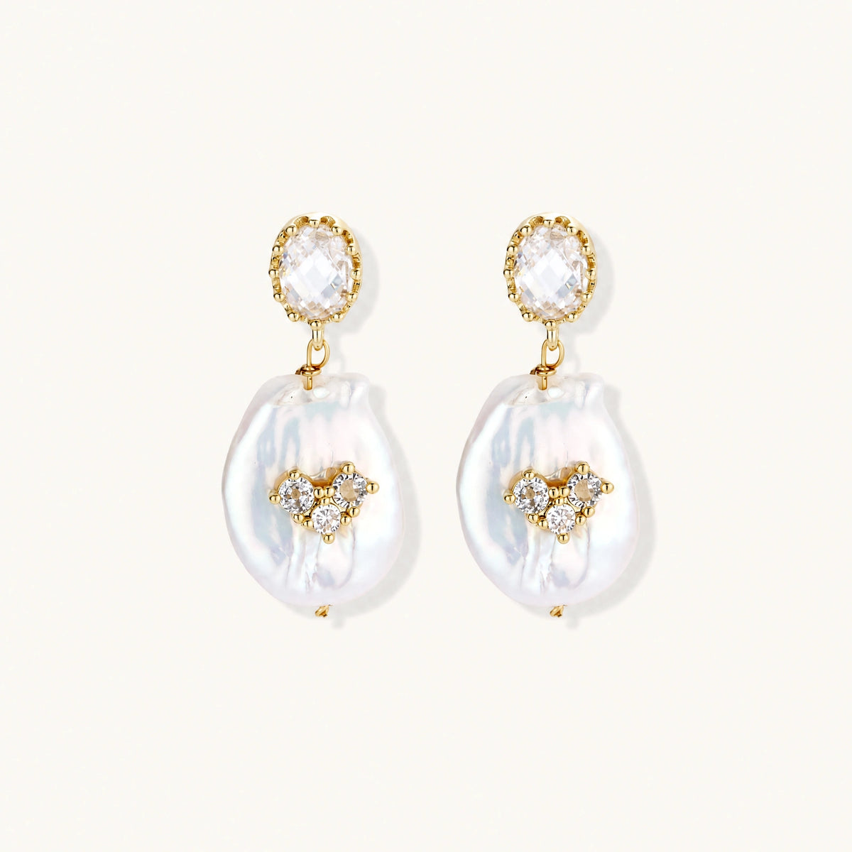 Gold-plated drop earrings with iridescent baroque pearls and crystal flower accents on cream background