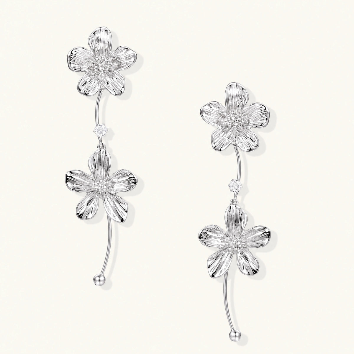 Silver-white asymmetric earrings with textured flower petals and crystal centers on cream background