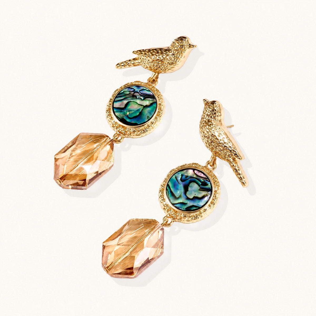 Gold-plated bird stud earrings with iridescent abalone shell medallions and champagne crystal drops on cream background