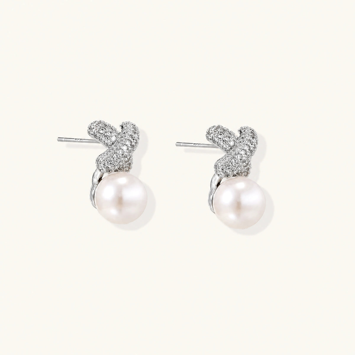 White gold plated studs featuring pavé X-shaped tops with freshwater pearl drops on cream background.