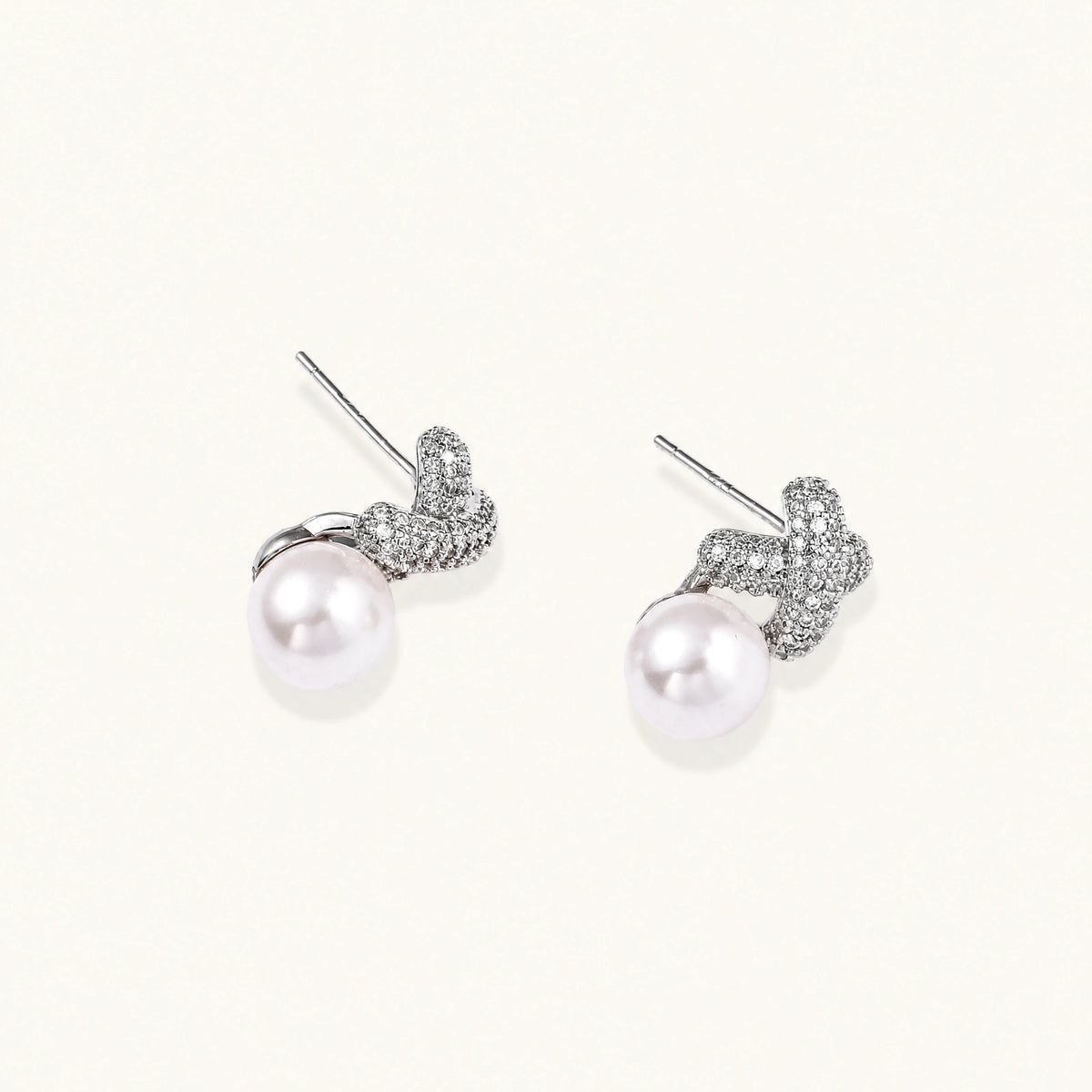 White gold plated studs featuring pavé X-shaped tops with freshwater pearl drops on cream background.