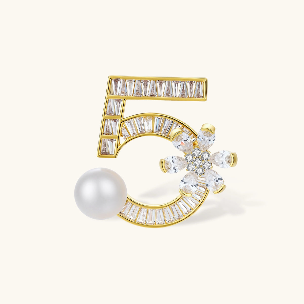 Gold-plated number 5 brooch with baguette zirconia, a crystal flower and a large white round bead on a cream background.
